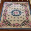 A cream dollhouse miniature rug with pink roses, blue flowers, and scrollwork border in a Victorian floral design.