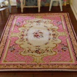 A pink, cream and burgundy dollhouse miniature rug with a central floral bouquet and scrollwork in a French country design.