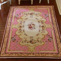 A pink, cream and burgundy dollhouse miniature rug with a central floral bouquet and scrollwork in a French country design.