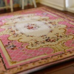 A pink, cream and burgundy dollhouse miniature rug with a central floral bouquet and scrollwork in a French country design.