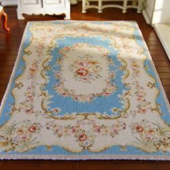 A cream dollhouse miniature rug with a central blue floral wreath surrounded by vines and scrollwork in shades of rosewood.