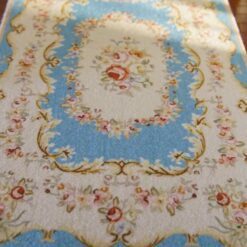 A cream dollhouse miniature rug with a central blue floral wreath surrounded by vines and scrollwork in shades of rosewood.