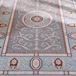 Soft blue and beige miniature rug with a floral medallion and intricate scrolling details.