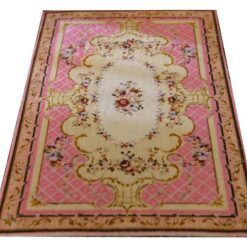 A pink, cream and burgundy dollhouse miniature rug with a central floral bouquet and scrollwork in a French country design.