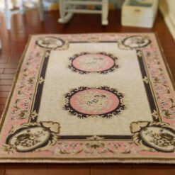 A cream dollhouse miniature rug with pink medallions, scrollwork borders, and floral accents in a French Aubusson design.