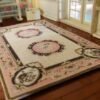 A cream dollhouse miniature rug with pink medallions, scrollwork borders, and floral accents in a French Aubusson design.