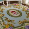 Victorian-style miniature rug with light blue background and floral roses, featuring scrolls and a central medallion.